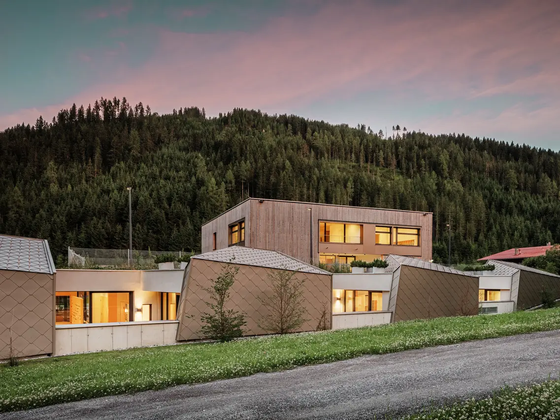L'immagine mostra il Centro educativo Gerlos al tramonto, con l'illuminazione interna. Con i suoi moderni rivestimenti in legno e i pannelli metallici a forma di diamante, l'edificio si inserisce armoniosamente nell'ambiente naturale della fitta foresta di conifere sullo sfondo. Il cielo è immerso nelle morbide tonalità della sera, creando un'atmosfera calma e invitante.