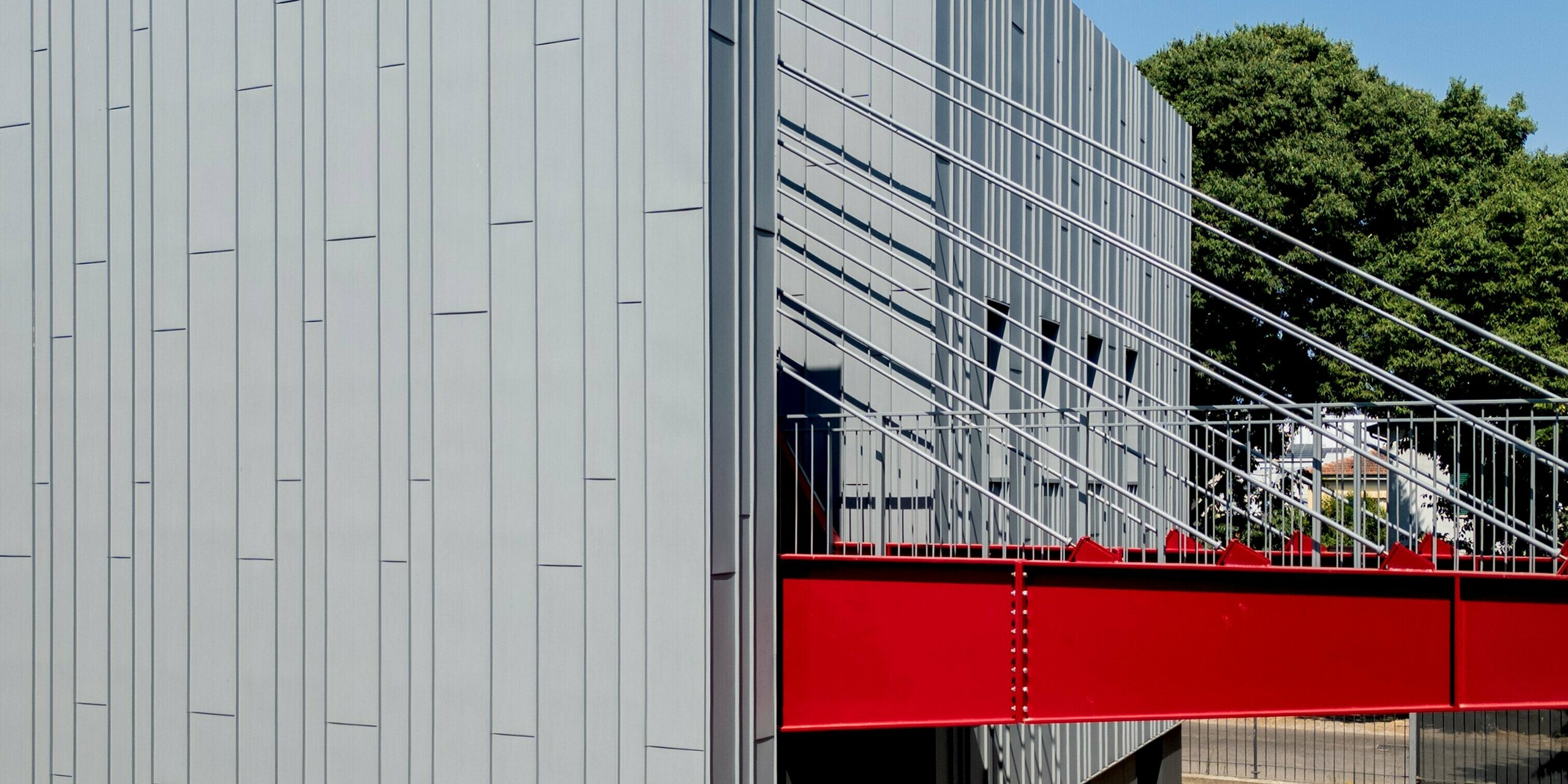 Ein kraftvoller Blickwinkel auf die Scuola Arte e Mestieri F. Bertazzoni in Suzzara zeigt eine spektakuläre, leuchtend rote Brückenkonstruktion, die sich zwischen zwei Gebäudeteilen spannt, sowie die vertikale PREFA-Aluminiumfassade in Patinagrau, die ihre ganze Präzision zeigt. Die makellose Verarbeitung von PREFALZ mit den charakteristischen Stehfalzbahnen unterstreicht die architektonische Stringenz des Gebäudes eindrucksvoll. Die gespannten Seile der Brücke werfen faszinierende Schattenspiele auf die Fassadenoberfläche. Die PREFA Aluminiumprodukte überzeugen hier mit dauerhafter Witterungsbeständigkeit, Formstabilität und zeitloser Ästhetik.