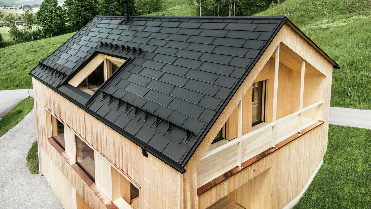 Vogelperspektive auf ein umweltfreundliches Holzhaus in Egg, eingebettet in grüne Wiesen, mit einem dunklen, langlebigen PREFA Solardach. Die schwarzen Dachplatten R.16 aus Aluminium harmonieren mit den Solarmodulen, die effizient in das Dach des Einfamilienhauses integriert sind, um nachhaltige Energie zu erzeugen. Das Gebäude mit seiner charakteristischen Holzfassade und modernen Architektur strahlt Natürlichkeit und zukunftsorientiertes Design aus, während es die Landschaft mit seiner fortschrittlichen Solartechnologie respektiert.