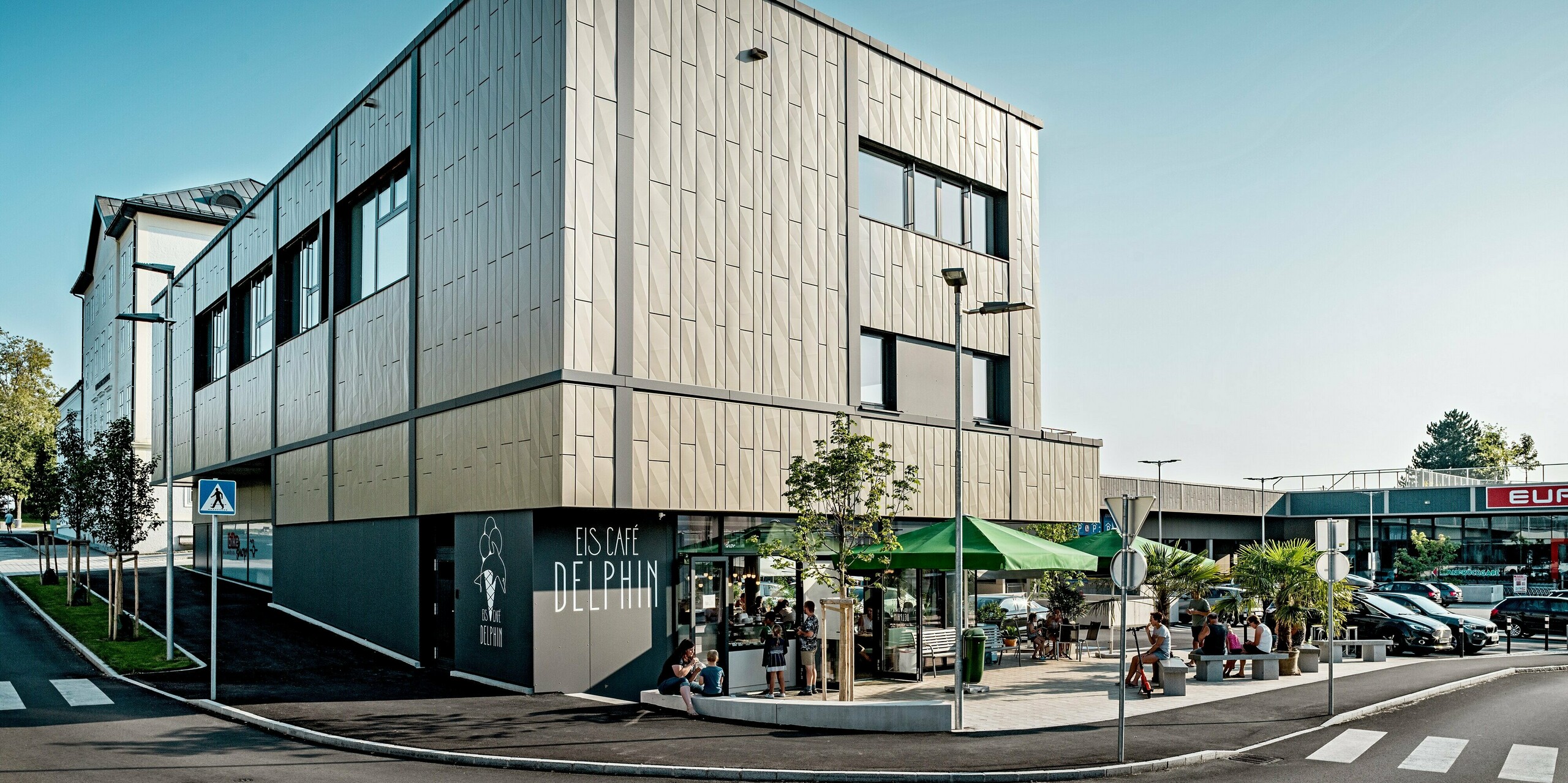 Das Bild zeigt die moderne Ecke des Einkaufszentrums in Rohrbach mit seiner eleganten Aluminiumfassade von PREFA. Die Fassade besteht aus Siding.X-Paneelen in den Farben Bronze und Anthrazit. Durch ihre feine Textur und das geometrische Design erzielen sie eine markante Wirkung. Vor dem Gebäude befindet sich das Eis Café Delphin mit einer einladenden Terrasse und Grünflächen, die für eine angenehme Atmosphäre sorgen. Die Fassade fügt sich harmonisch in die Umgebung ein und verbindet Funktionalität mit modernem Design.