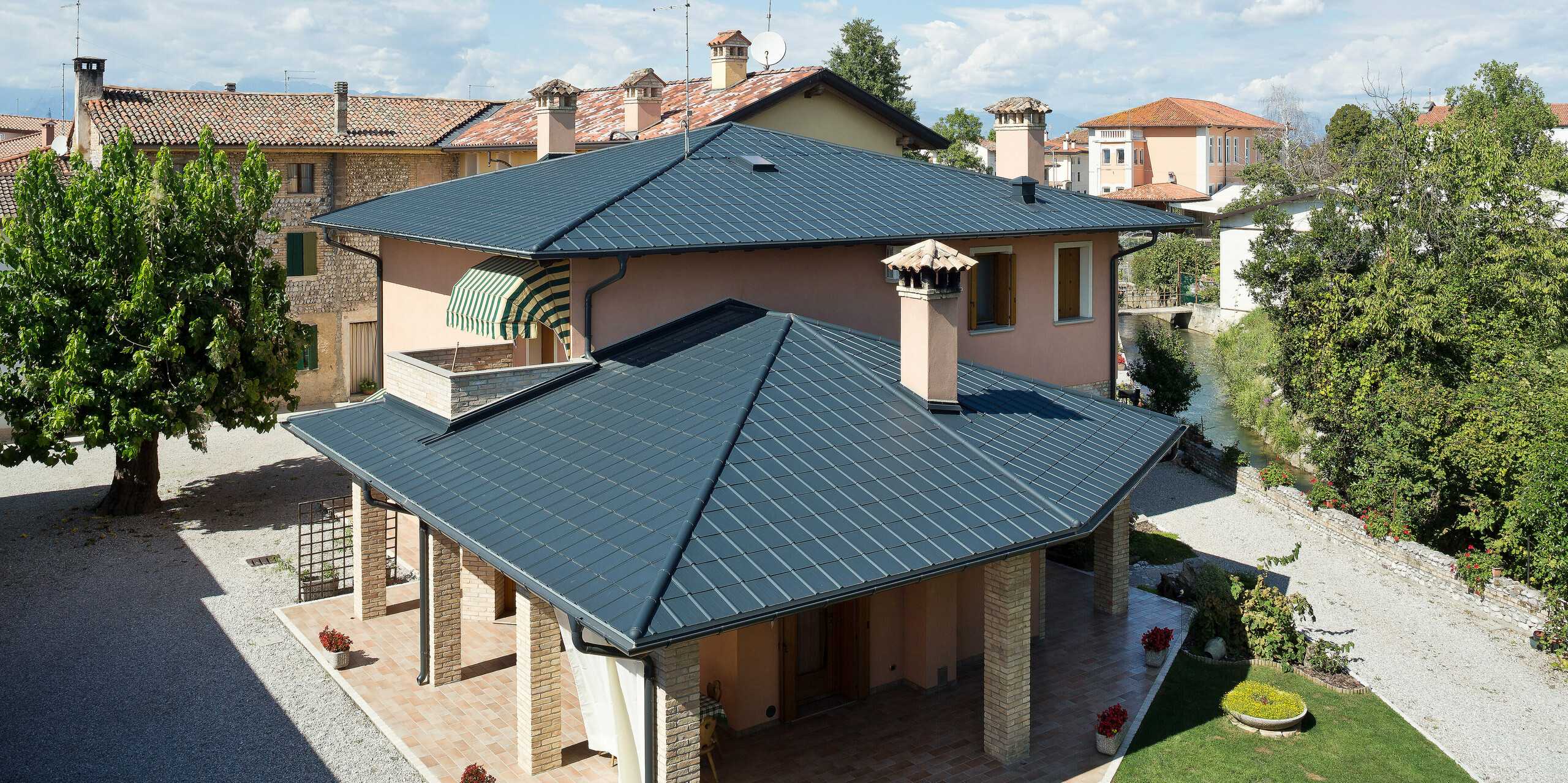 Elegantes Dachdesign in in San Lorenzo di Arzene: Die Villa Beroia setzt auf PREFA P.10 Dachplatten in Anthrazit. So vereint das Gebäude italienische Tradition mit einem modernen Aluminiumdach. In der Umgebung befinden sich Schotterwege, Bäume und Sträucher sowie ein kleiner Fluss, der direkt am Haus vorbeifließt.
