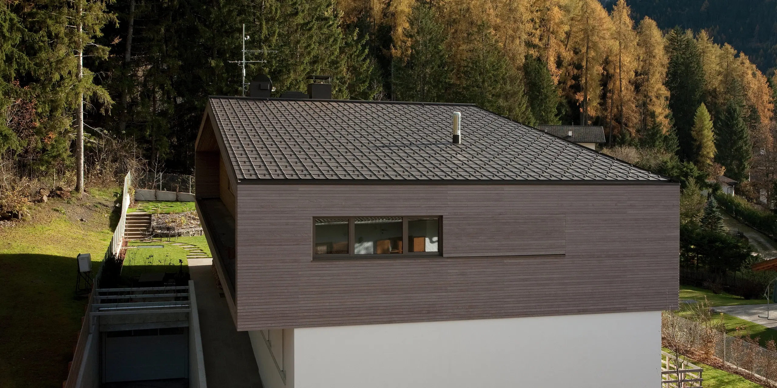 Moderne Architektur mit PREFA Dachplatten in P.10 Braun, umgeben von einem naturnahen Garten und Waldlandschaft. Der minimalistische Baukörper mit Holzfassade fügt sich harmonisch in die Umgebung ein.