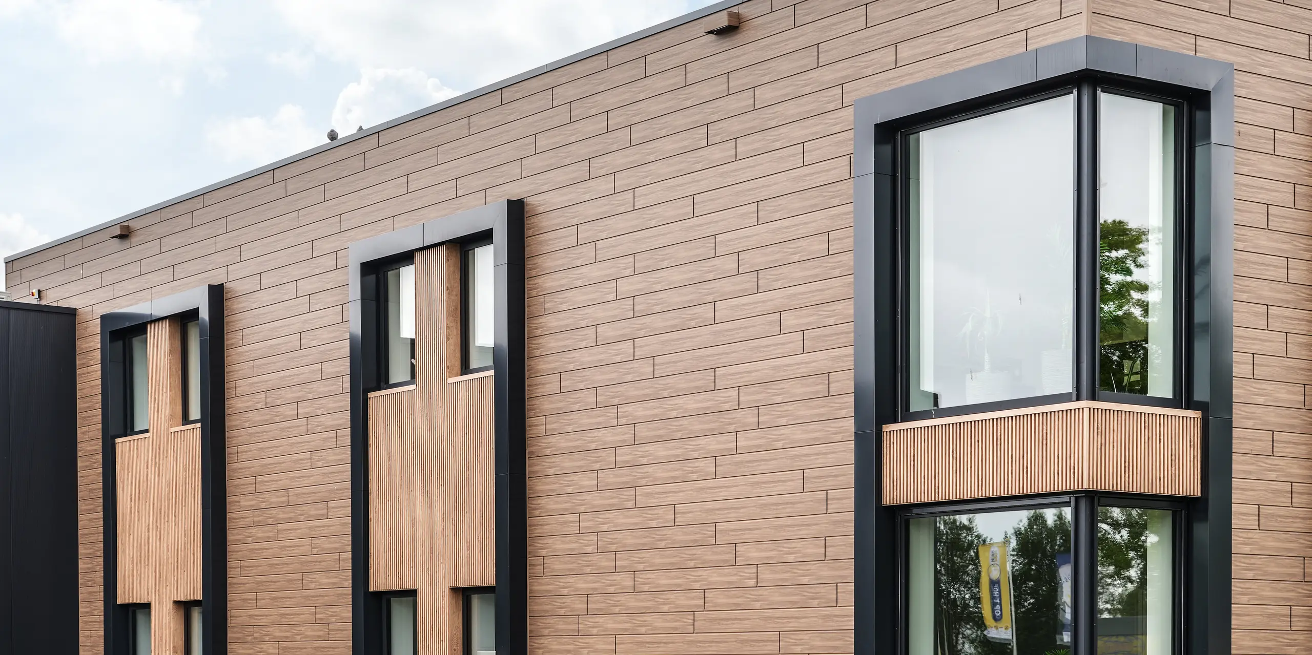 Detailansicht des Firmengebäudes der Dokter Schoonmaakorganisatie in Wolvega mit präzise gestalteter PREFA Fassade in Walnussbraun. Die horizontal verlegten Sidings in Holzoptik verleihen der Gebäudehülle eine warme, natürliche Wirkung, während die tief eingeschnittenen, dunkel gerahmten Fenster die moderne Architektur betonen. Klare Linien, starke Kontraste und hochwertige Materialien schaffen einen markanten Gesamteindruck.
