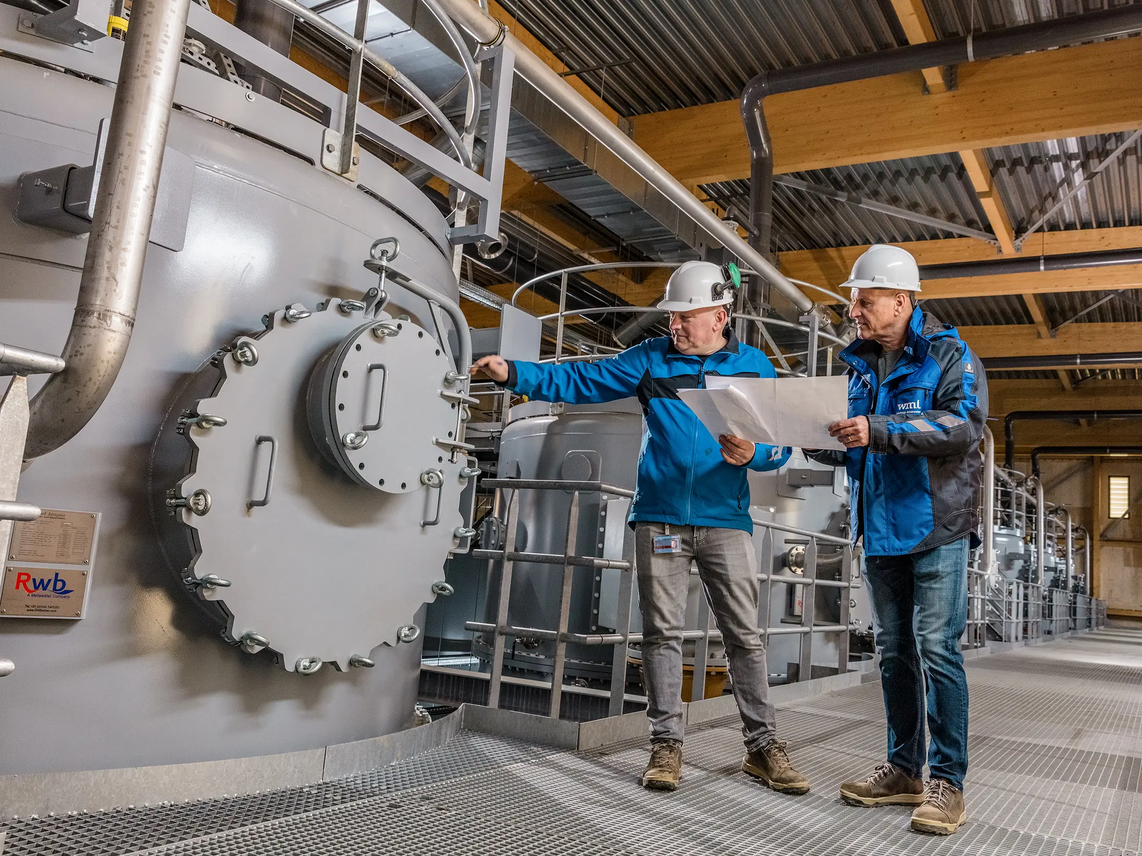 Im modernen Trinkwasserpumpwerk von WML in Lottum arbeiten zwei Fachkräfte zwischen großdimensionierten Filteranlagen, Rohrleitungen und präziser Technik unter einer markanten Holzkonstruktion. Im Fokus stehen robuste, zylindrische Behälter und die industrielle Klarheit des Raums. Die leistungsfähige Architektur setzt außen auf langlebige PREFA Aluminiumfassaden, die mit geringem Gewicht, hoher Witterungsbeständigkeit und nachhaltiger Recyclingfähigkeit überzeugen. Das Zusammenspiel aus Hightech, Holz und innovativen Materialien macht das Infrastrukturgebäude zu einem zukunftsweisenden Eyecatcher.
