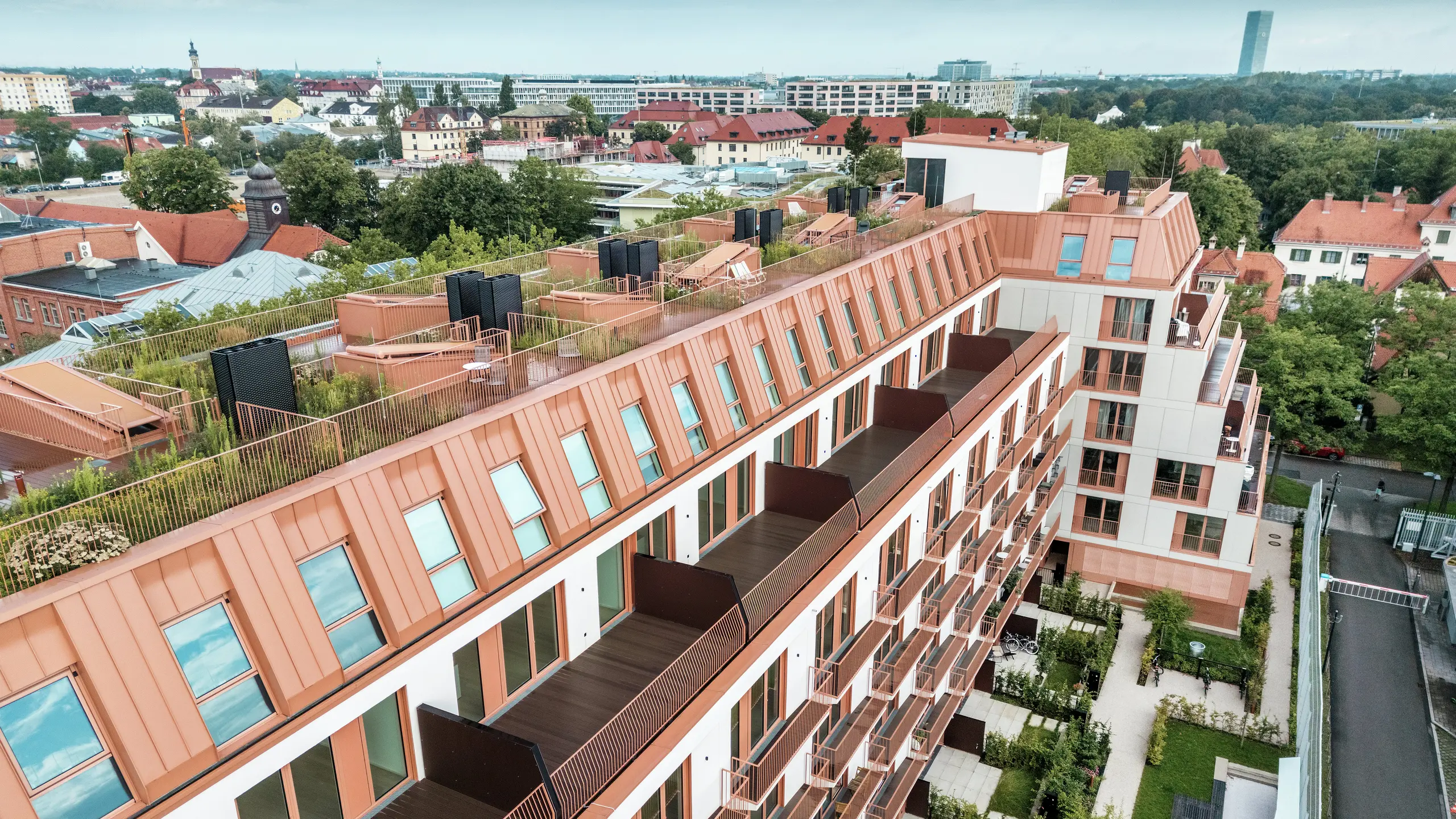 Vista aerea del progetto residenziale Van B a Monaco di Baviera con il suggestivo tetto in alluminio PREFALZ nel colore speciale IGP Orange HDP di PREFA.  Ampi balconi, terrazze verdi sul tetto e un preciso design della facciata sottolineano il concetto architettonico ben studiato. Ideale per uno sviluppo urbano sostenibile e per una vita urbana con un'alta qualità di vita e un'estetica accattivante.
