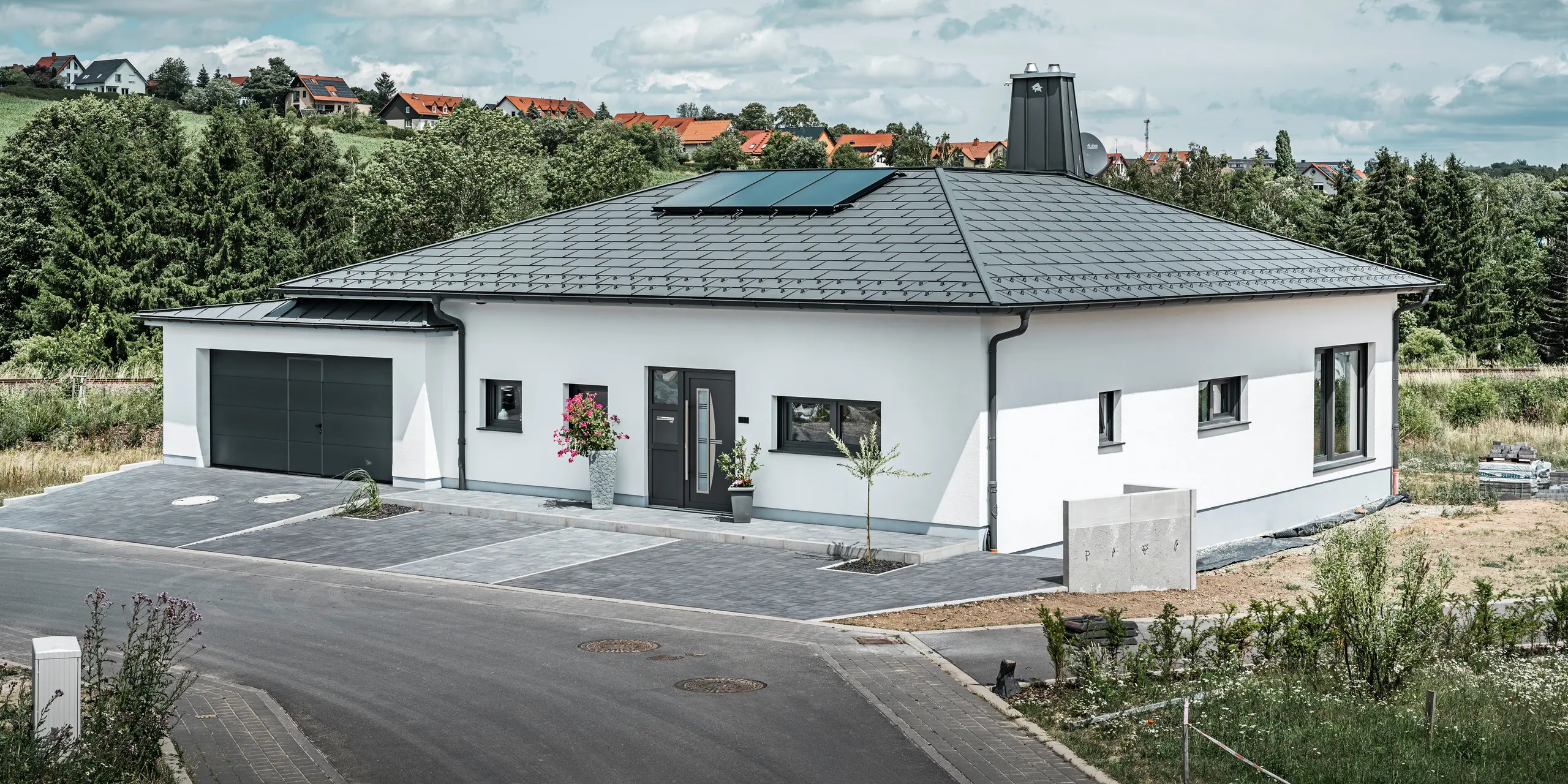 Elegante casa unifamiliare a Ilmenau, in Turingia, con un tetto PREFA di alta qualità nel colore P.10 grigio scuro, dotato della robusta tegola R.16. Il garage si armonizza nel colore e nello stile grazie all'utilizzo di PREFALZ. Il punto di forza della proprietà è l'atrio dal design funzionale e il camino decorato con il toro PREFA, che sottolineano il design moderno e l'innovativa tecnologia dei tetti PREFA. I pannelli solari si integrano perfettamente nel concetto sostenibile della casa. Il concetto globale ben studiato del bungalow è completato dal sistema di drenaggio del tetto PREFA, anch'esso in grigio scuro.