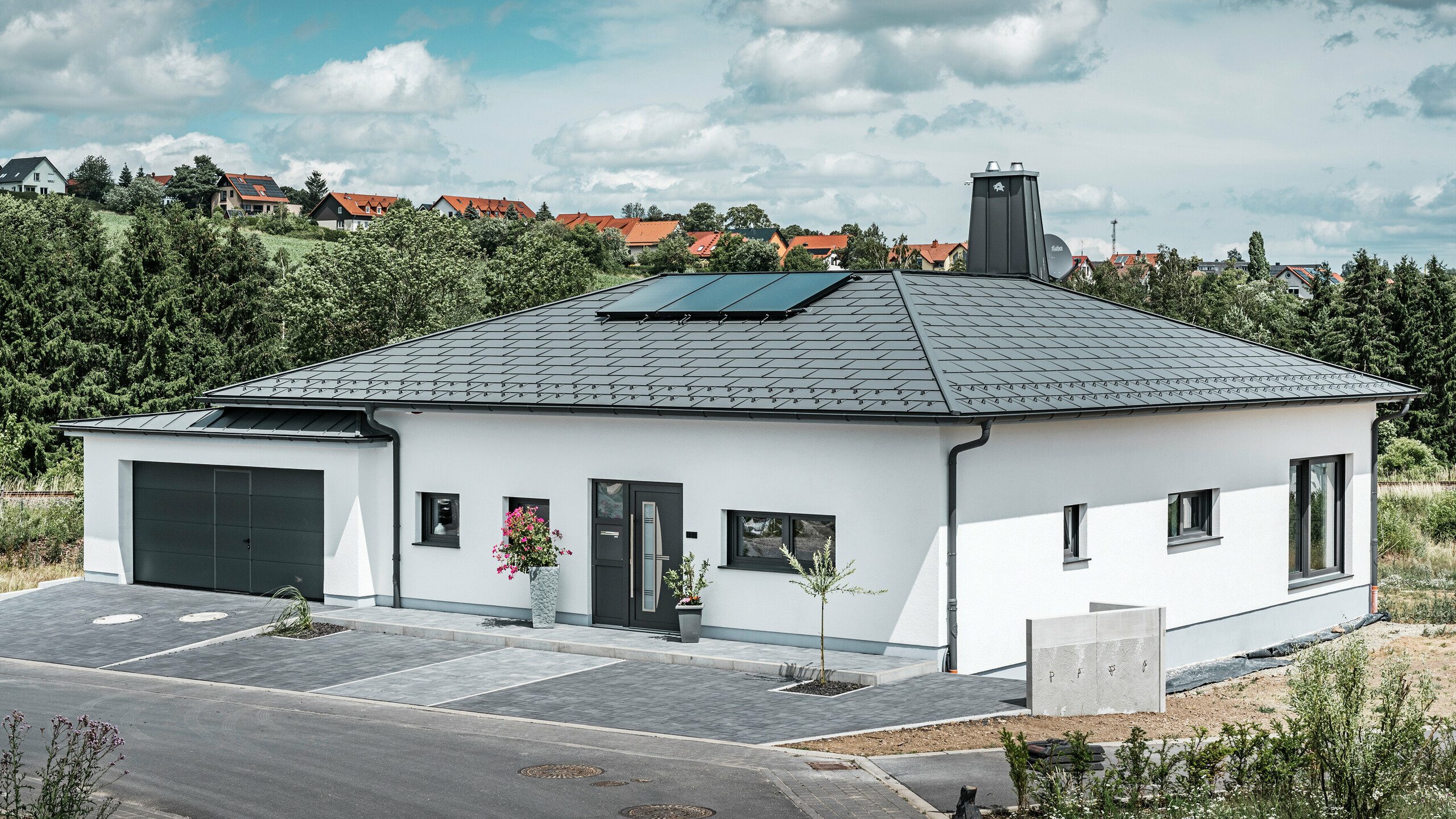 Elegantes Einfamilienhaus in Ilmenau, Thüringen, mit einem hochwertigen PREFA Dach in der Farbe P.10 Dunkelgrau, ausgestattet mit der robusten Dachplatte R.16. Die Garage harmoniert in Farbe und Stil durch die Verwendung von PREFALZ. Der Blickfang des Anwesens ist das funktional gestaltete Atrium und der mit dem PREFA Stier verzierte Schornstein, welcher das moderne Design und die innovative Dachtechnologie von PREFA unterstreichen. Die Solarpaneele fügen sich nahtlos in das nachhaltige Konzept des Hauses ein. Abgerundet wird das durchdachte Gesamtkonzept des Bungalows durch das PREFA Dachentwässerungssytem, welches ebenfalls in Dunkelgrau gehalten ist.