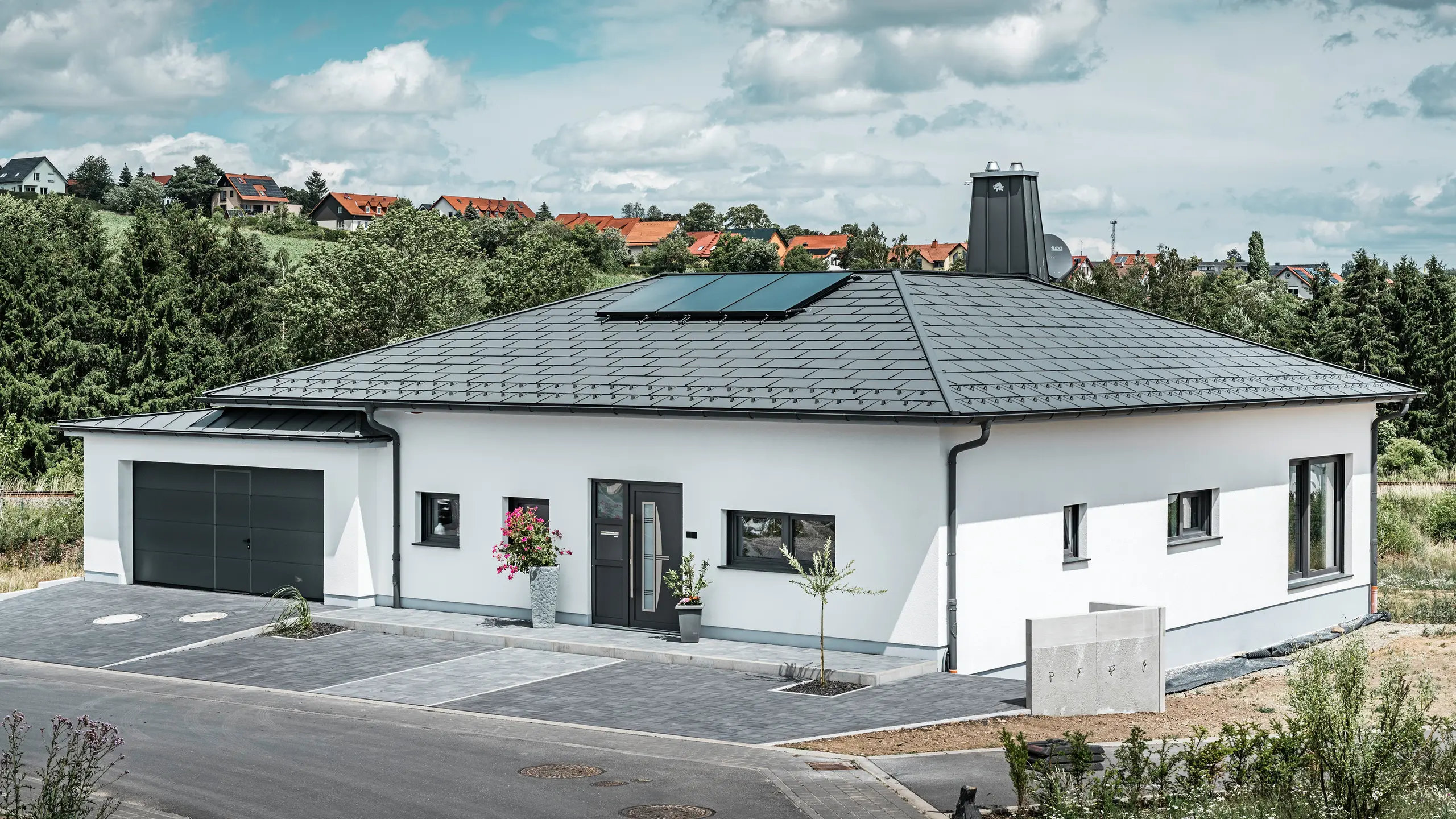 Elegante casa unifamiliare a Ilmenau, in Turingia, con un tetto PREFA di alta qualità nel colore P.10 grigio scuro, dotato della robusta tegola R.16. Il garage si armonizza nel colore e nello stile grazie all'utilizzo di PREFALZ. Il punto di forza della proprietà è l'atrio dal design funzionale e il camino decorato con il toro PREFA, che sottolineano il design moderno e l'innovativa tecnologia dei tetti PREFA. I pannelli solari si integrano perfettamente nel concetto sostenibile della casa. Il concetto globale ben studiato del bungalow è completato dal sistema di drenaggio del tetto PREFA, anch'esso in grigio scuro.