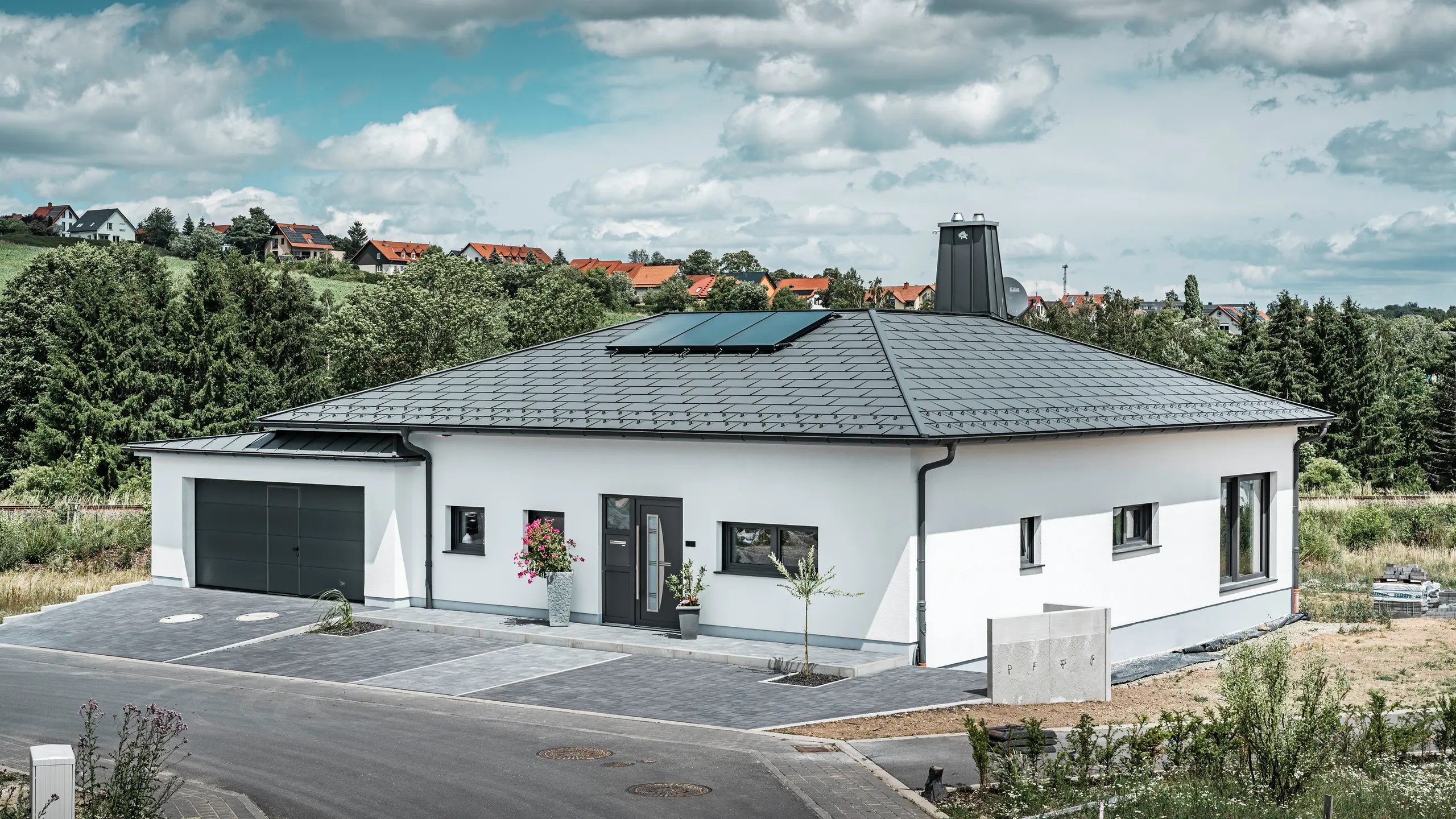 Elegante casa unifamiliare a Ilmenau, in Turingia, con un tetto PREFA di alta qualità nel colore P.10 grigio scuro, dotato della robusta tegola R.16. Il garage si armonizza nel colore e nello stile grazie all'utilizzo di PREFALZ. Il punto di forza della proprietà è l'atrio dal design funzionale e il camino decorato con il toro PREFA, che sottolineano il design moderno e l'innovativa tecnologia dei tetti PREFA. I pannelli solari si integrano perfettamente nel concetto sostenibile della casa. Il concetto globale ben studiato del bungalow è completato dal sistema di drenaggio del tetto PREFA, anch'esso in grigio scuro.