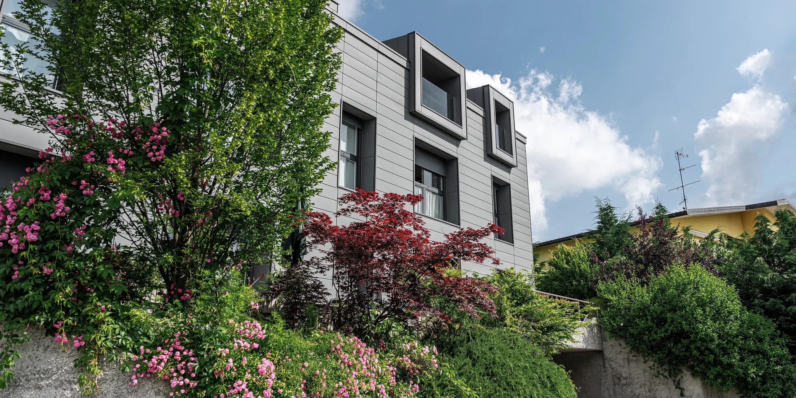 Moderno complesso residenziale a Faloppio, Italia, con elegante facciata realizzata con doga PREFA. Le finestre a sbalzo offrono un elemento architettonico unico, mentre le piante e i fiori circostanti sottolineano la bellezza naturale dell'edificio. Il rivestimento in alluminio PREFA, di lunga durata, garantisce una soluzione esteticamente gradevole e funzionale in qualsiasi condizione atmosferica.