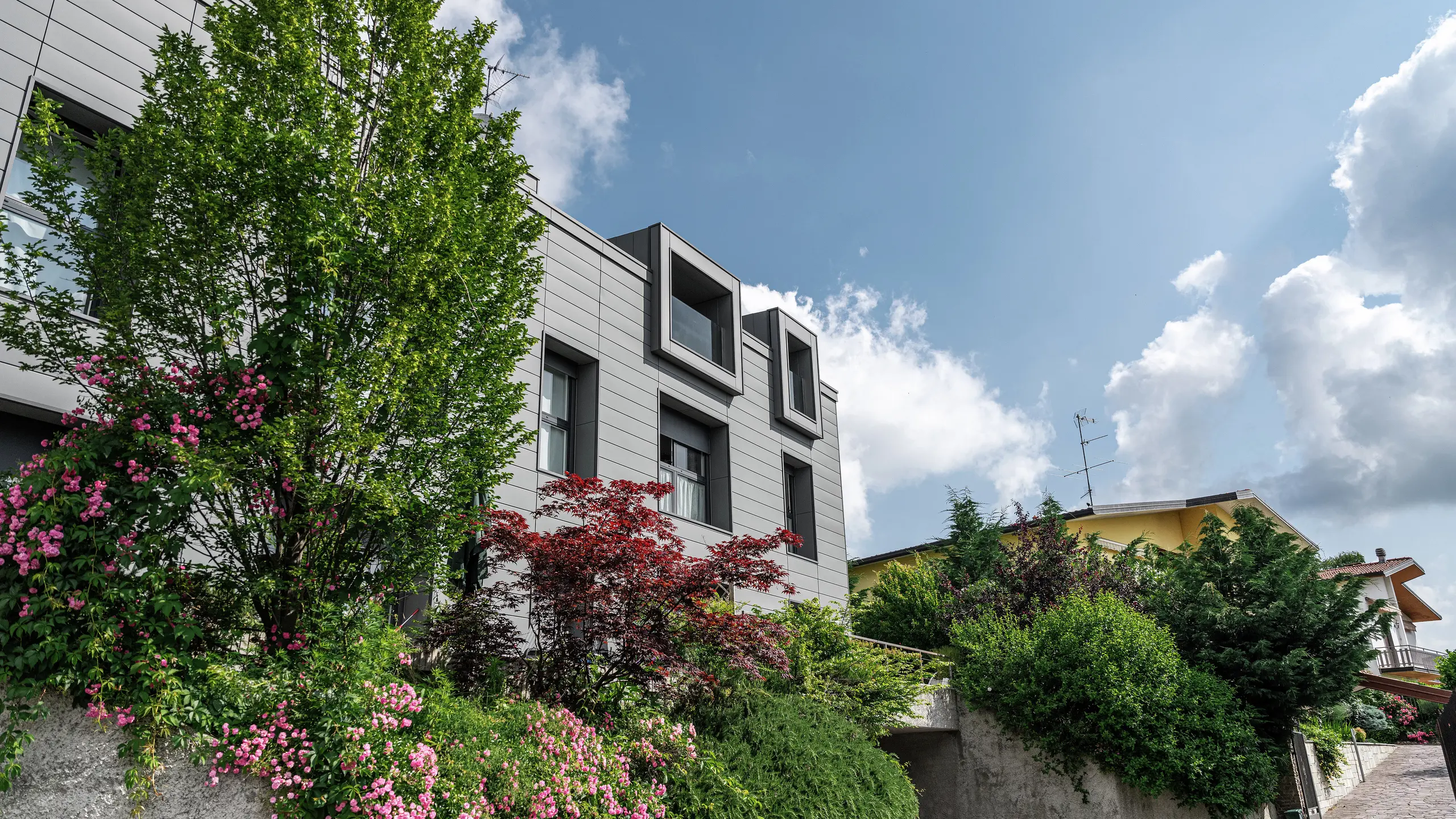 Moderno complesso residenziale a Faloppio, Italia, con elegante facciata realizzata con doga PREFA. Le finestre a sbalzo offrono un elemento architettonico unico, mentre le piante e i fiori circostanti sottolineano la bellezza naturale dell'edificio. Il rivestimento in alluminio PREFA, di lunga durata, garantisce una soluzione esteticamente gradevole e funzionale in qualsiasi condizione atmosferica.
