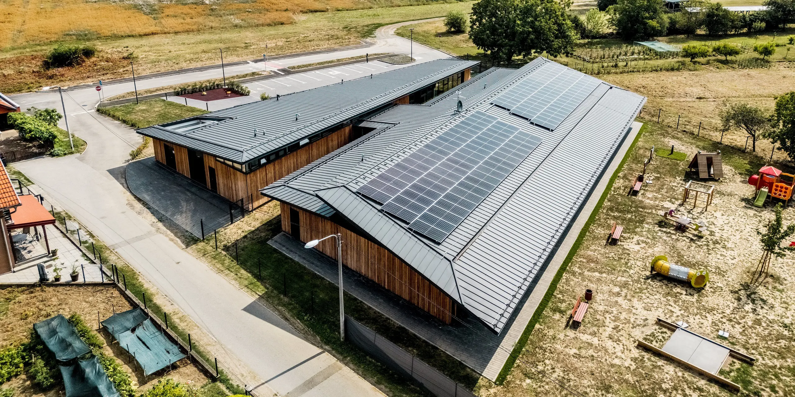 Luftaufnahme des Kindergartens Svarozic mit markanter PREFALZ Dachlandschaft in P.10 Anthrazit, großflächiger Photovoltaikanlage und natürlicher Holzfassade. Das moderne Gebäude liegt eingebettet in eine grüne Umgebung mit Spielplatz und Freiflächen. Das PREFA Aluminiumdach überzeugt durch geringes Gewicht, hohe Widerstandsfähigkeit und dauerhaften Schutz bei klarer, zeitloser Gestaltung.