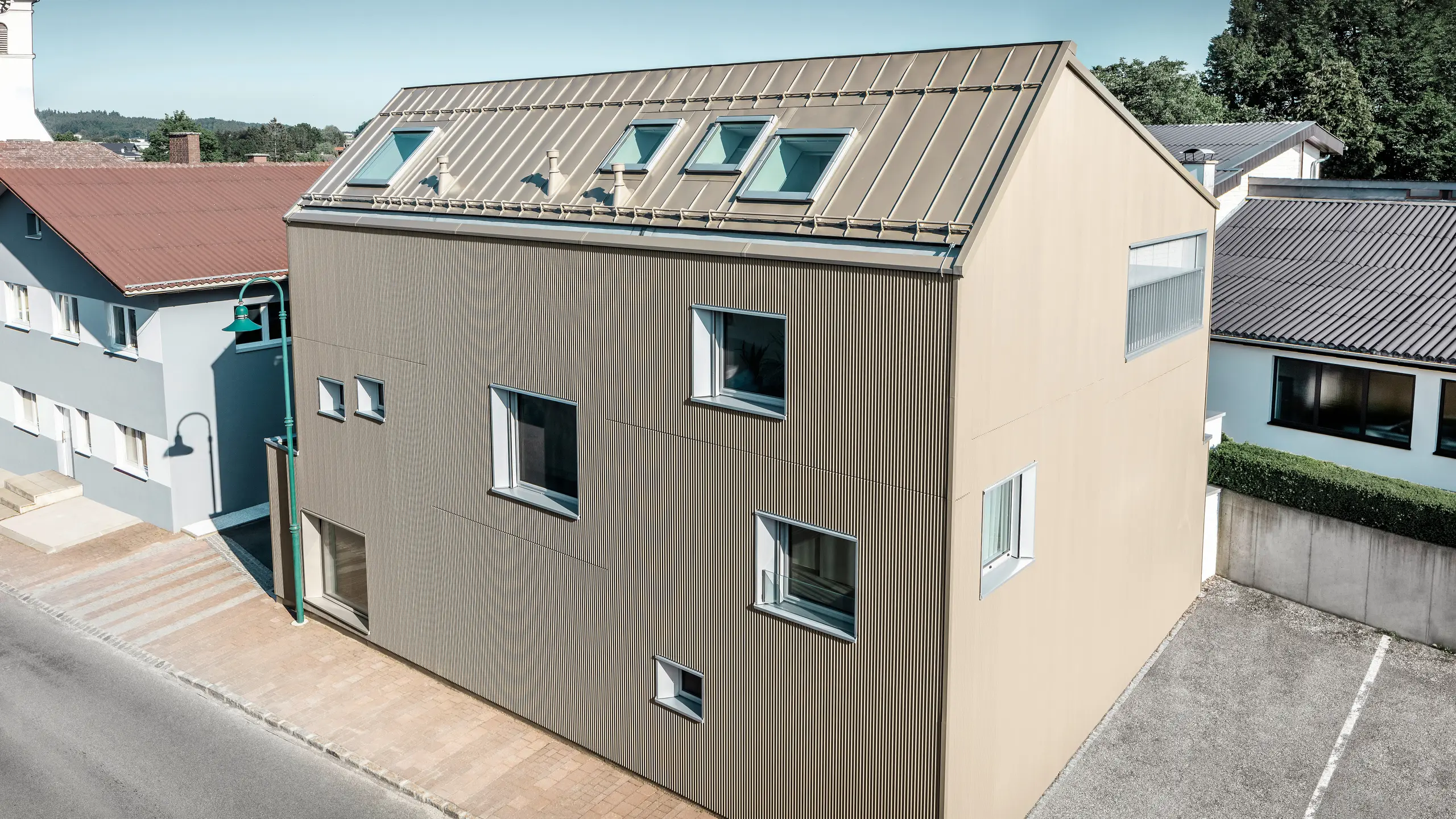 Vogelperspektive auf ein Einfamilienhaus in Frankenburg, Österreich, das hervorsticht durch seine PREFA Fassade in Form des Zackenprofils und einem PREFALZ-Dach, beide in abgestimmtem Bronze-Farbton. Die Fassade zeigt eine markante Textur, die das natürliche Licht einfängt und für ein lebendiges Schattenspiel sorgt. Mehrere Dachfenster lassen natürliches Licht ins Innere und unterstreichen die funktionale und ästhetische Integration moderner Architekturelemente. Das Haus fügt sich durch sein zeitgemäßes Design harmonisch in die typisch österreichische Dorf-Umgebung ein und setzt gleichzeitig einen kontrastreichen Akzent gegenüber den umliegenden traditionellen Gebäuden.