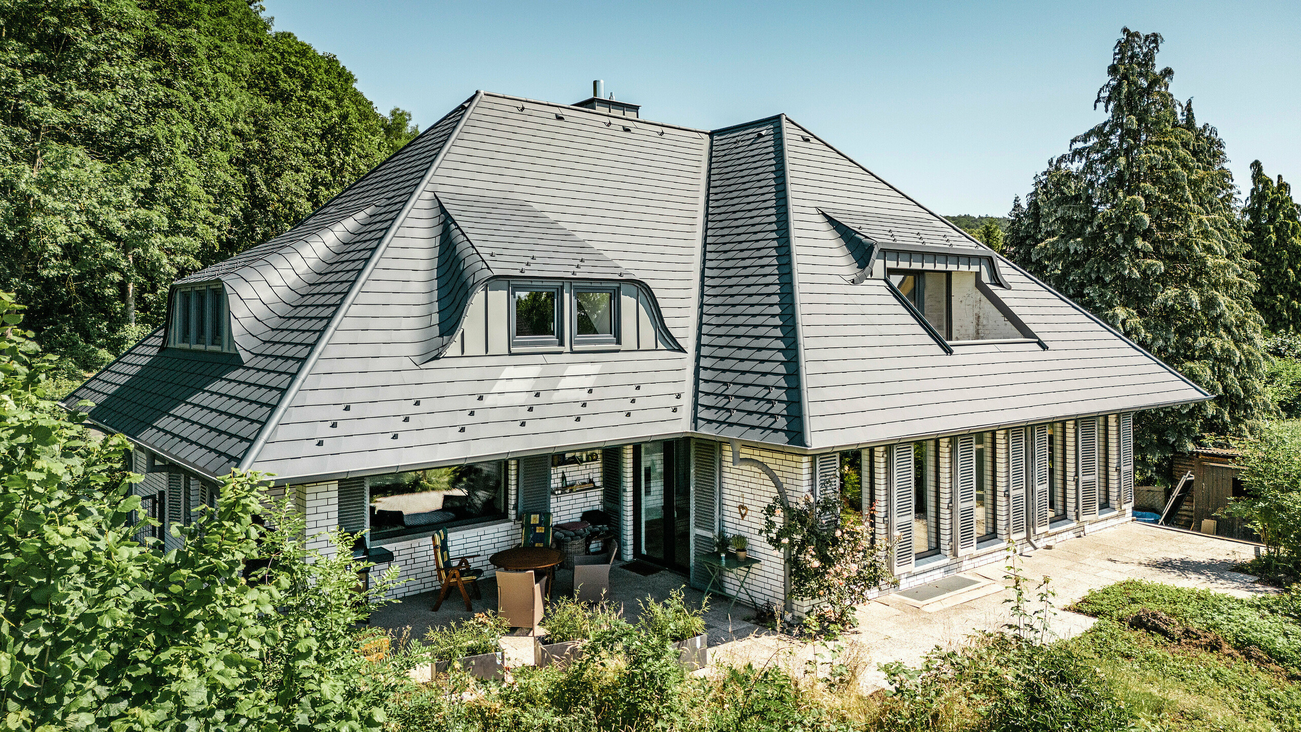Moderna casa unifamiliare a Einbeck, Germania, con tegole Scandola PREFA in P.10 antracite. Le tegole PREFA in alluminio, di lunga durata, sono note per la loro qualità e resistenza. Il design del tetto dell'edificio sottolinea l'architettura individuale con abbaini a capanna, che vengono messi in risalto dalla flessibilità delle tegole PREFA. Anche il sistema di drenaggio del tetto, costituito dal tubo pluviale PREFA e dalla grondaia semicircolare, è in antracite. La casa unifamiliare è circondata da un ampio giardino ben curato e da una terrazza. Dietro la casa si trova un bosco che, insieme al cielo azzurro, crea un'atmosfera naturale.