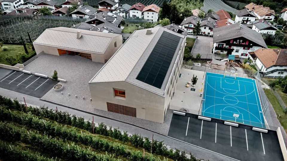 Veduta aerea del centro educativo di Riffian in Alto Adige, con scuola materna, scuola elementare e area sportiva. Le scaglie 44 × 44 PREFA in P.10 bronze formano una copertura in alluminio precisa e durevole e si integrano armoniosamente nel contesto alpino. L’architettura moderna offre una protezione resistente alle intemperie e un design pulito nei dettagli.