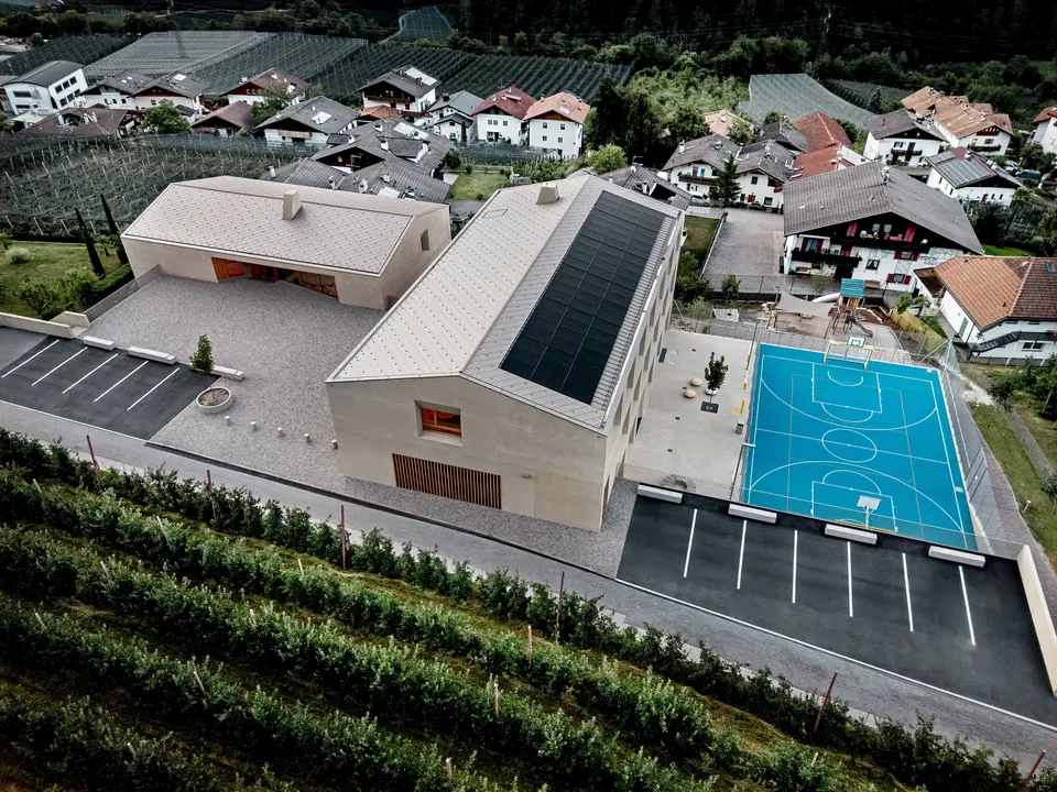 Veduta aerea del centro educativo di Riffian in Alto Adige, con scuola materna, scuola elementare e area sportiva. Le scaglie 44 × 44 PREFA in P.10 bronze formano una copertura in alluminio precisa e durevole e si integrano armoniosamente nel contesto alpino. L’architettura moderna offre una protezione resistente alle intemperie e un design pulito nei dettagli.