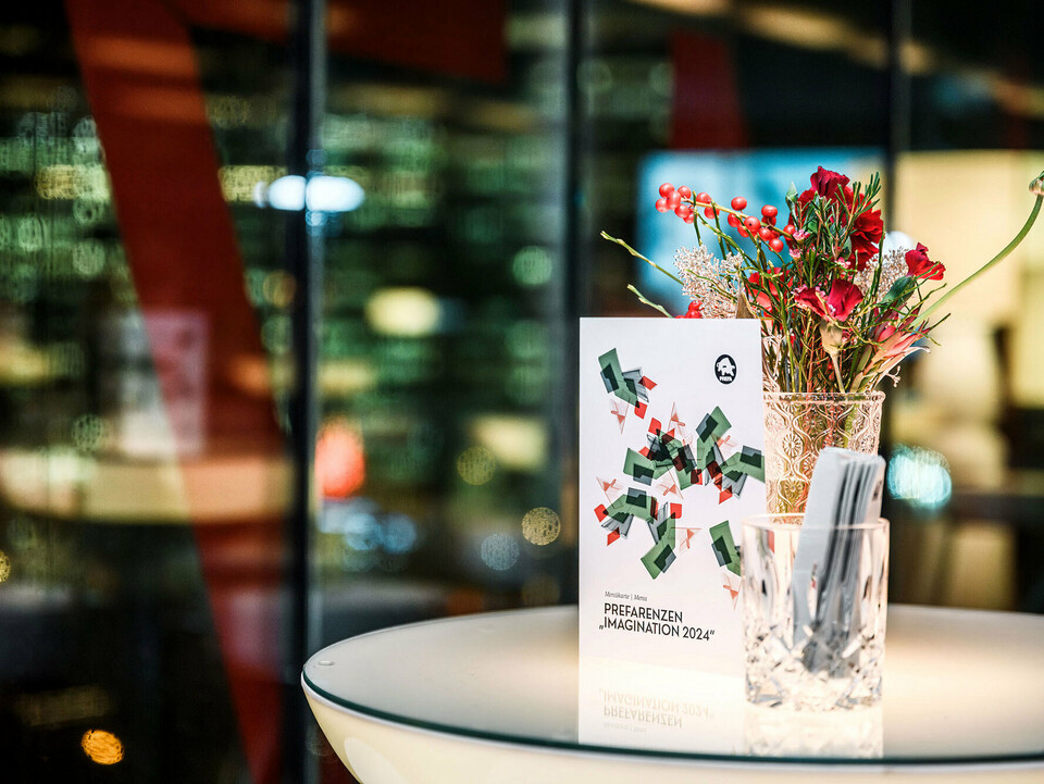 Scatto dell'invito all'evento PREFARENZEN 2024 posizionato su un tavolo davanti a una composizione floreale in una brocca di vetro su uno sfondo sfocato.