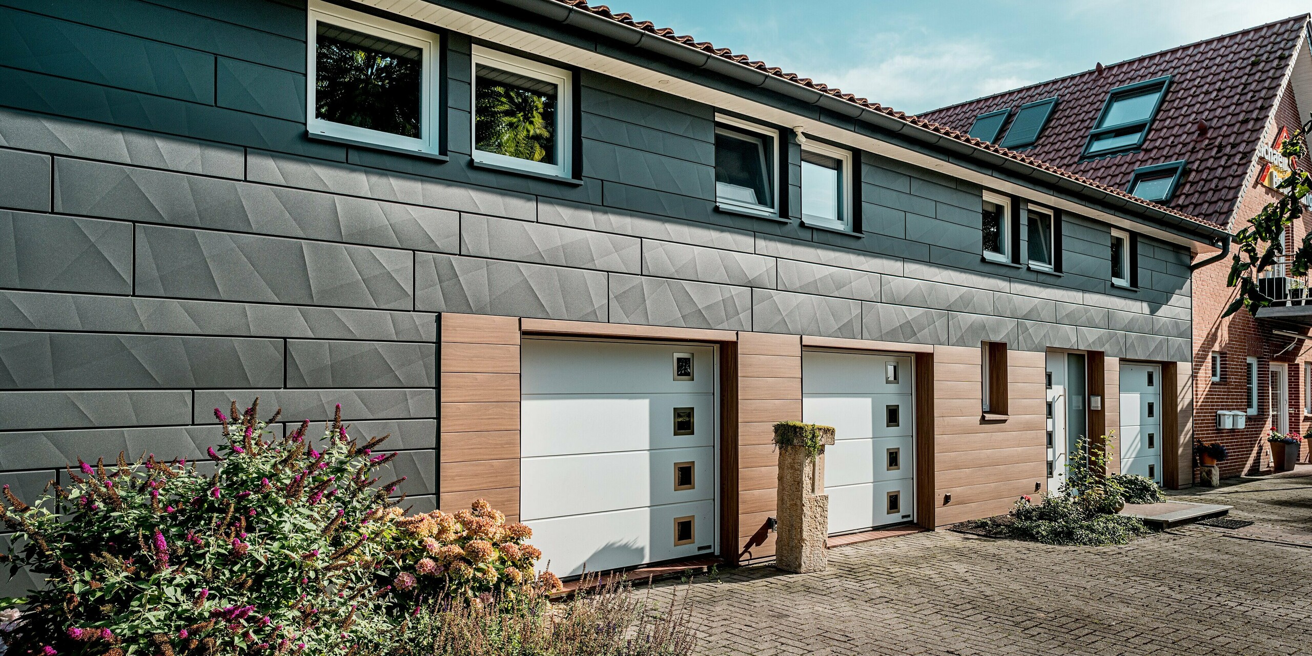 Schrägansicht eines sanierten Einfamilienhauses mit moderner PREFA Aluminiumfassade und klarer, horizontaler Struktur. Besonders ins Auge fallen die großflächigen PREFA Siding.X Paneele in P.10 Dunkelgrau mit ihren markanten Längs- und Querkantungen, die ein lebendiges Spiel aus Licht und Schatten erzeugen. Im unteren Fassadenbereich setzen PREFA Siding Paneele in Walnussbraun mit edler Holzoptik warme, einladende Akzente rund um Garagentore und Eingänge. Die weißen Tore und Fensterrahmen schaffen starke Kontraste. Die langlebigen, pflegeleichten und witterungsbeständigen PREFA Aluminiumpaneele machen diese Fassadensanierung zu einem ausdrucksstarken Beispiel moderner, nachhaltiger Architektur.