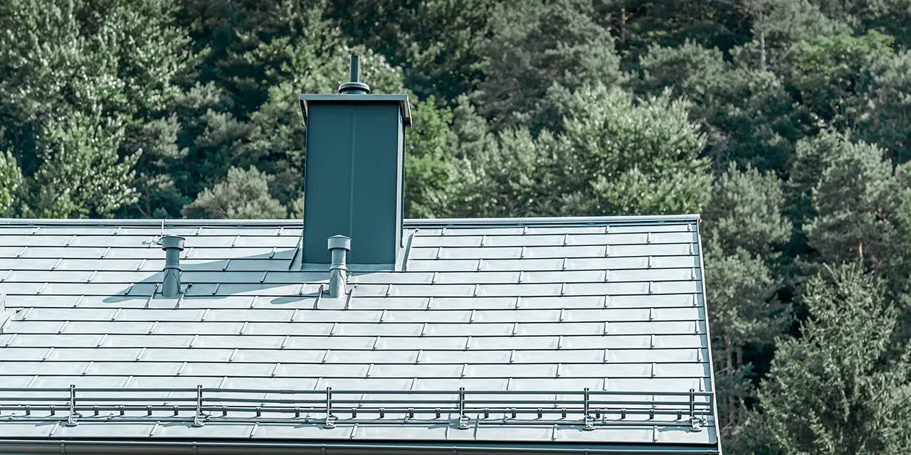 Immagine dettagliata di un tetto a due falde, rivestito con la tegola R.16 PREFA in grigio pietra con sistema paraneve, conversa per camino, grondaia e tubo di ventilazione, sempre PREFA