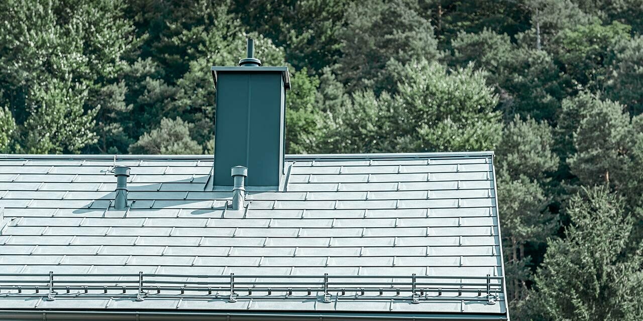 Immagine dettagliata di un tetto a due falde, rivestito con la tegola R.16 PREFA in grigio pietra con sistema paraneve, conversa per camino, grondaia e tubo di ventilazione, sempre PREFA