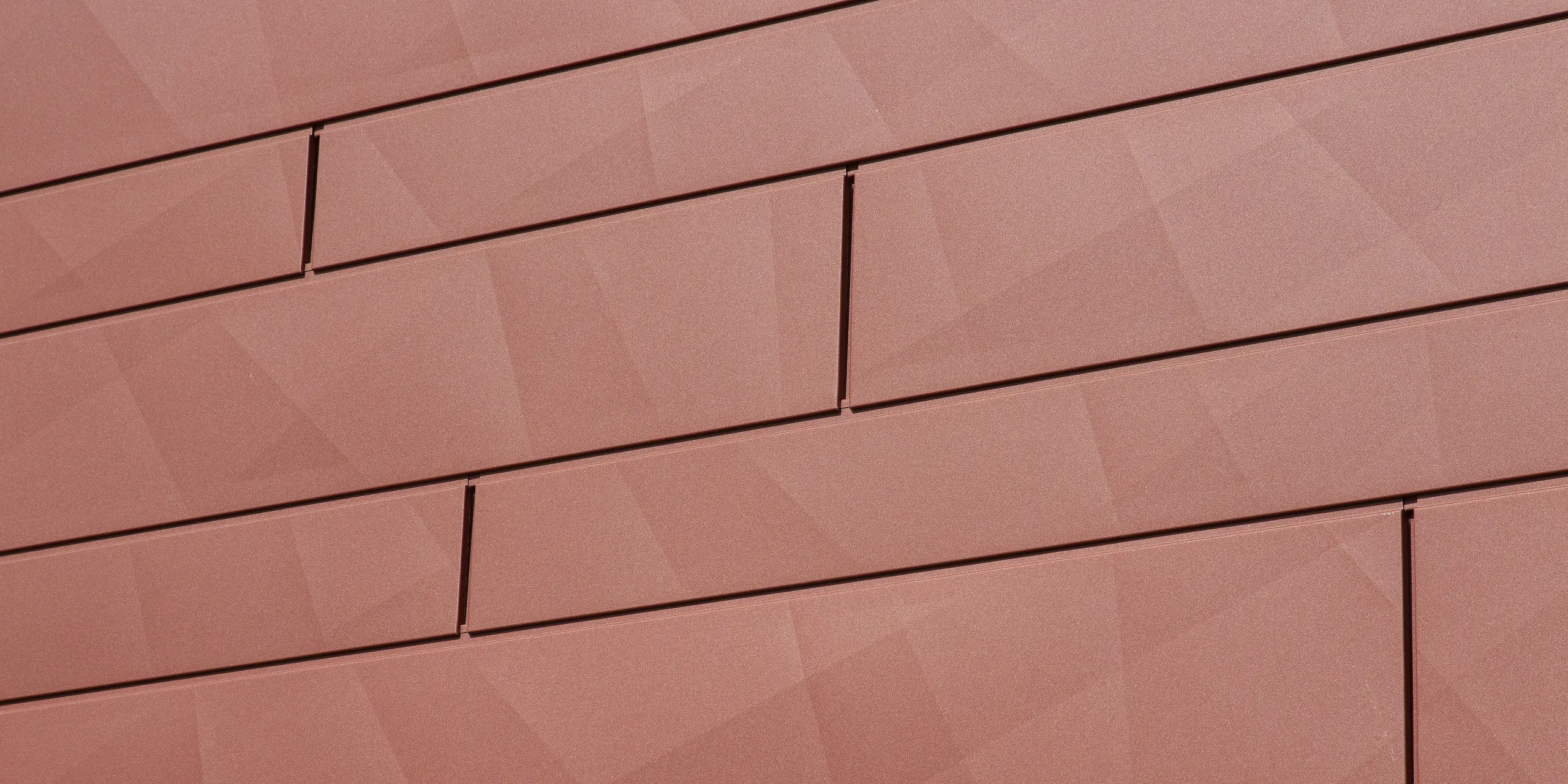 Detailaufnahme der PREFA Fassadenpaneele FX.12 in Oxydrot: Die prägnante Kantung erzeugt eine spannende geometrische Struktur, die das Licht je nach Tageszeit anders reflektiert. Die hochwertige Pulverbeschichtung in P.10 Qualität sorgt für dauerhafte Farbbrillanz und höchste Widerstandsfähigkeit – ideal für langlebige, wartungsfreie Fassadensanierungen.
