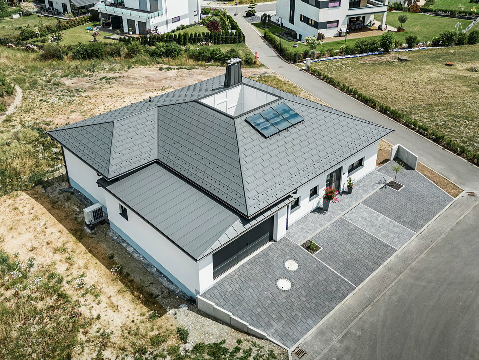 Moderne Architektur trifft Funktionalität: Ein Bungalow in Ilmenau, Thüringen, mit einem beeindruckenden PREFA Aluminiumdach bestehend aus der Dachplatte R.16 in P.10 Dunkelgrau. Die Dachlandschaft zeigt ein zentrales Atrium und einen markanten Schornstein mit dem charakteristischen PREFA Stier, ergänzt durch ein langlebiges PREFA Dachentwässerungssystem mit Ablaufrohr und Hängerinne. Die Garage fügt sich nahtlos in das Gesamtbild ein, verkleidet mit PREFALZ in passendem Dunkelgrau.