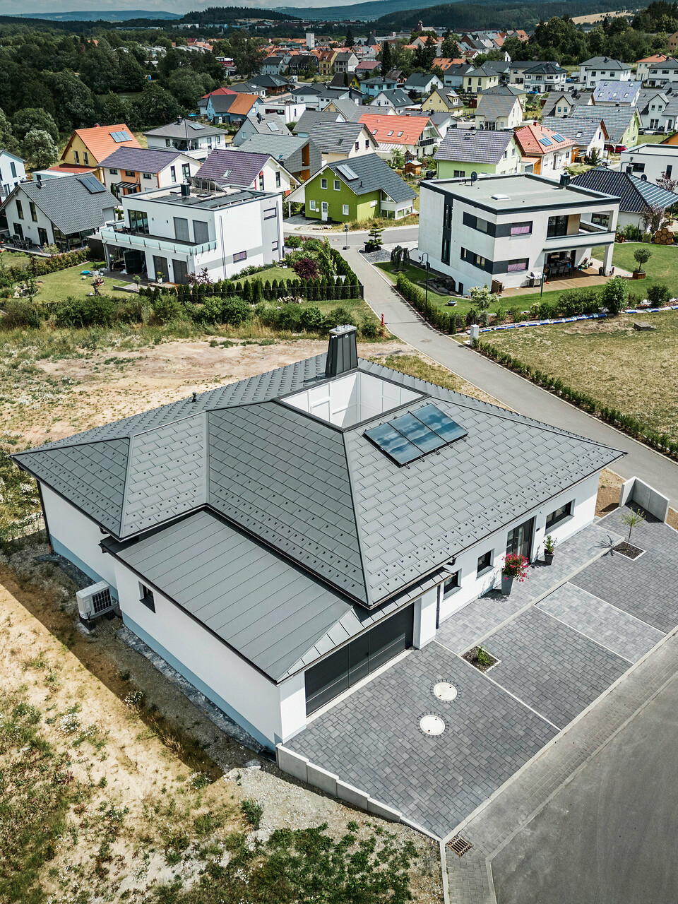 Moderne Architektur trifft Funktionalität: Ein Bungalow in Ilmenau, Thüringen, mit einem beeindruckenden PREFA Aluminiumdach bestehend aus der Dachplatte R.16 in P.10 Dunkelgrau. Die Dachlandschaft zeigt ein zentrales Atrium und einen markanten Schornstein mit dem charakteristischen PREFA Stier, ergänzt durch ein langlebiges PREFA Dachentwässerungssystem mit Ablaufrohr und Hängerinne. Die Garage fügt sich nahtlos in das Gesamtbild ein, verkleidet mit PREFALZ in passendem Dunkelgrau.