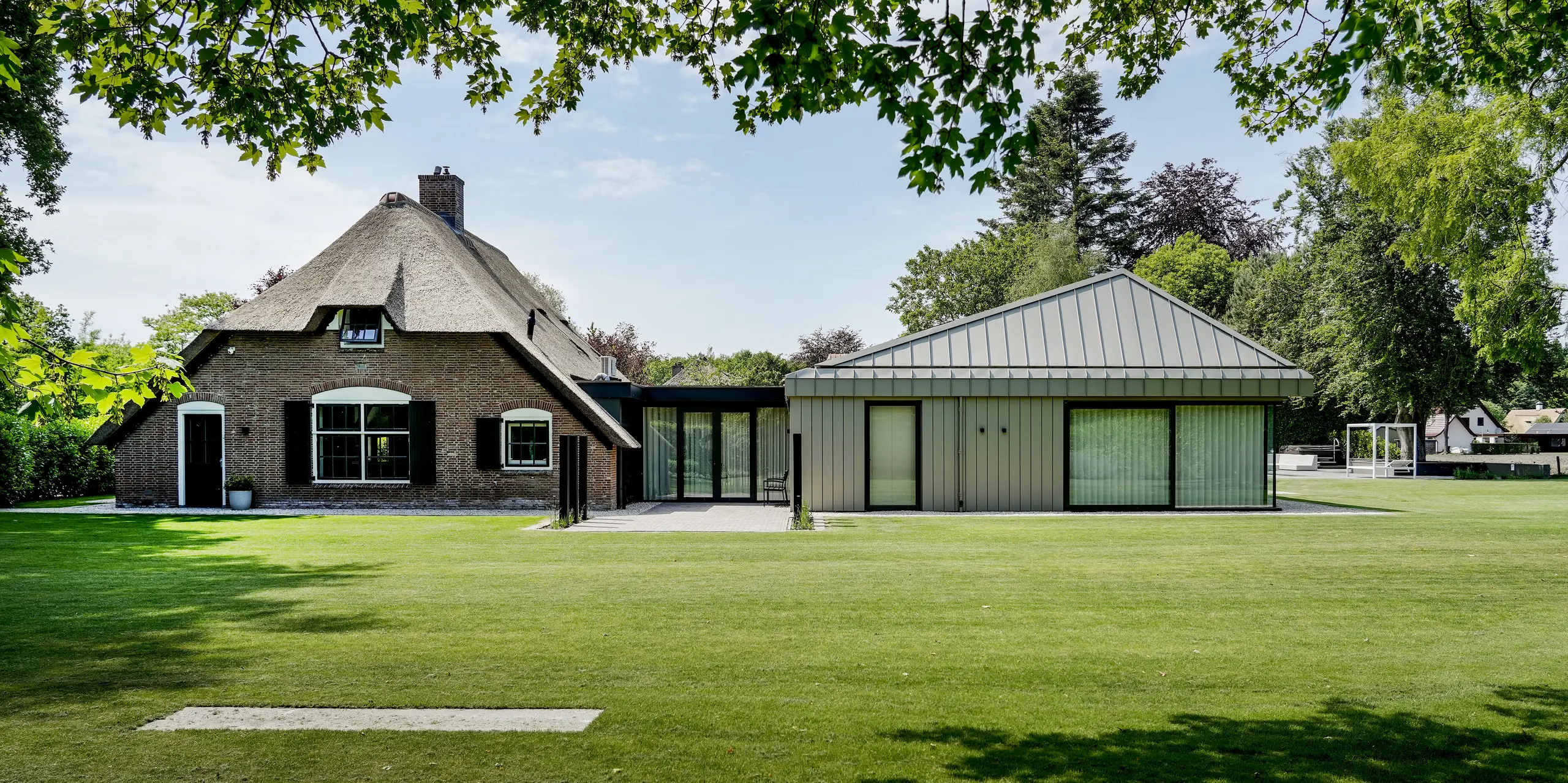 Weitläufige Gartenansicht eines Einfamilienhauses in Putten, in dem traditionelles Reetdach und moderner Anbau mit PREFALZ Aluminium eindrucksvoll aufeinandertreffen. Die klare Dachform, die durchgehende Aluminiumfassade und großzügige Glasflächen setzen starke architektonische Akzente. PREFA Aluminium überzeugt durch langlebige Qualität, zeitlose Ästhetik und eine harmonische Verbindung von Alt und Neu.