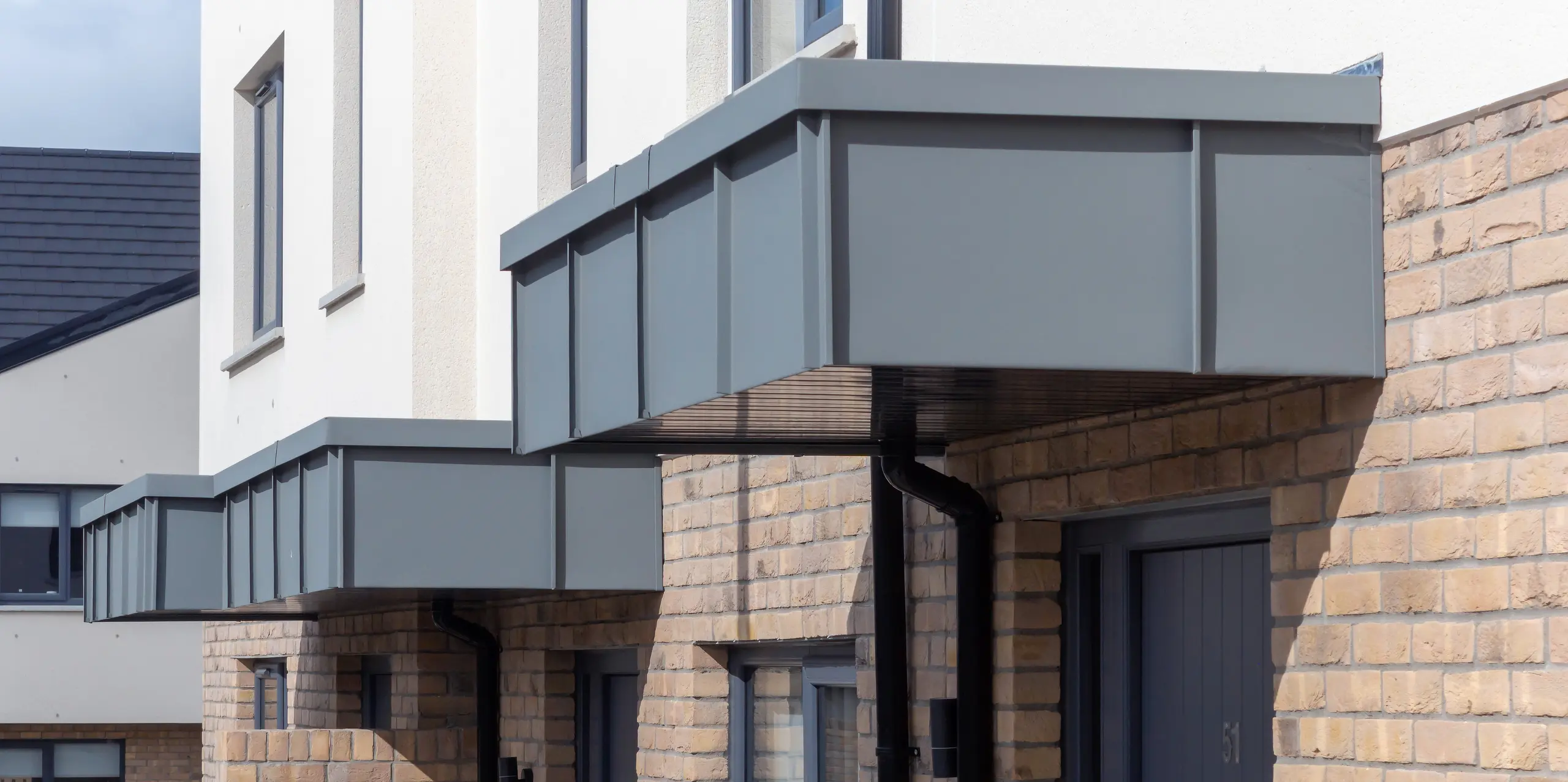 Das Bild zeigt die markanten Eingangsvordächer der Reihenhäuser in Churchfields, die vollständig mit PREFALZ in P.10 Hellgrau verkleidet sind. Die klaren, geradlinigen Aluminiumflächen setzen einen modernen Akzent über dem warmen Backsteinmauerwerk und verleihen der Anlage ein einheitliches, hochwertiges Erscheinungsbild. Die robuste PREFA Oberfläche ist witterungsbeständig, farbstabil und nahezu wartungsfrei – ideal für langlebige Wohnbauten. Die Detailaufnahme betont die präzise Verarbeitung und die harmonische Integration der Aluminiumverkleidung in die Architektur.
