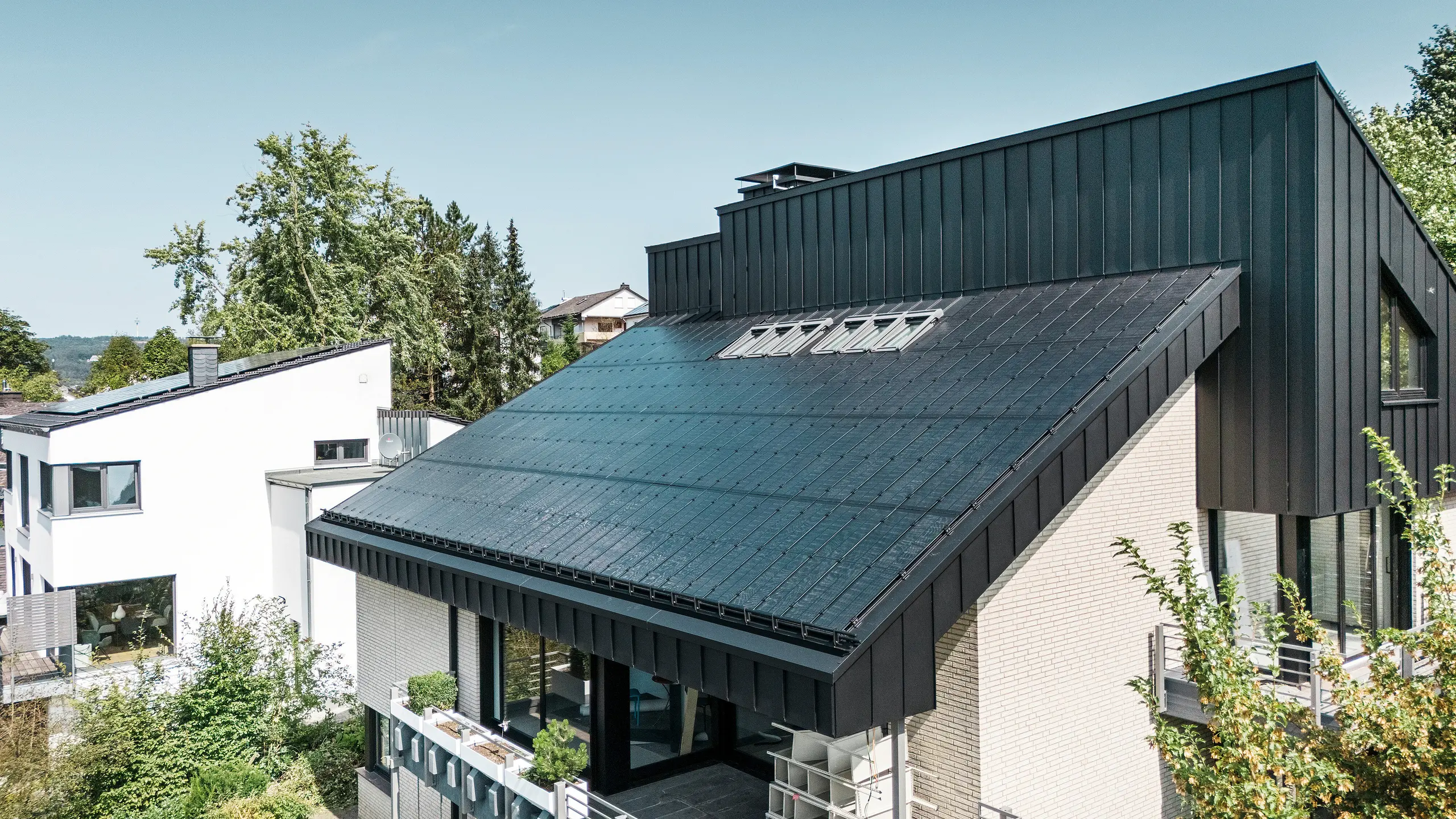 Architettura moderna di una casa unifamiliare a Meschede, in Germania, con un tetto in alluminio PREFALZ in P.10 nero e moduli fotovoltaici vetro/vetro integrati. Il tetto a una falda sfalsato combina un design senza tempo con una tecnologia fotovoltaica sostenibile. Gli innovativi moduli fotovoltaici PREFALZ forniscono una potenza di 18 kWp. I moduli senza cornice e l'elegante rivestimento in alluminio di facciata e tetto formano un'unità estetica e funzionale che si integra perfettamente nel verde ambiente residenziale.
