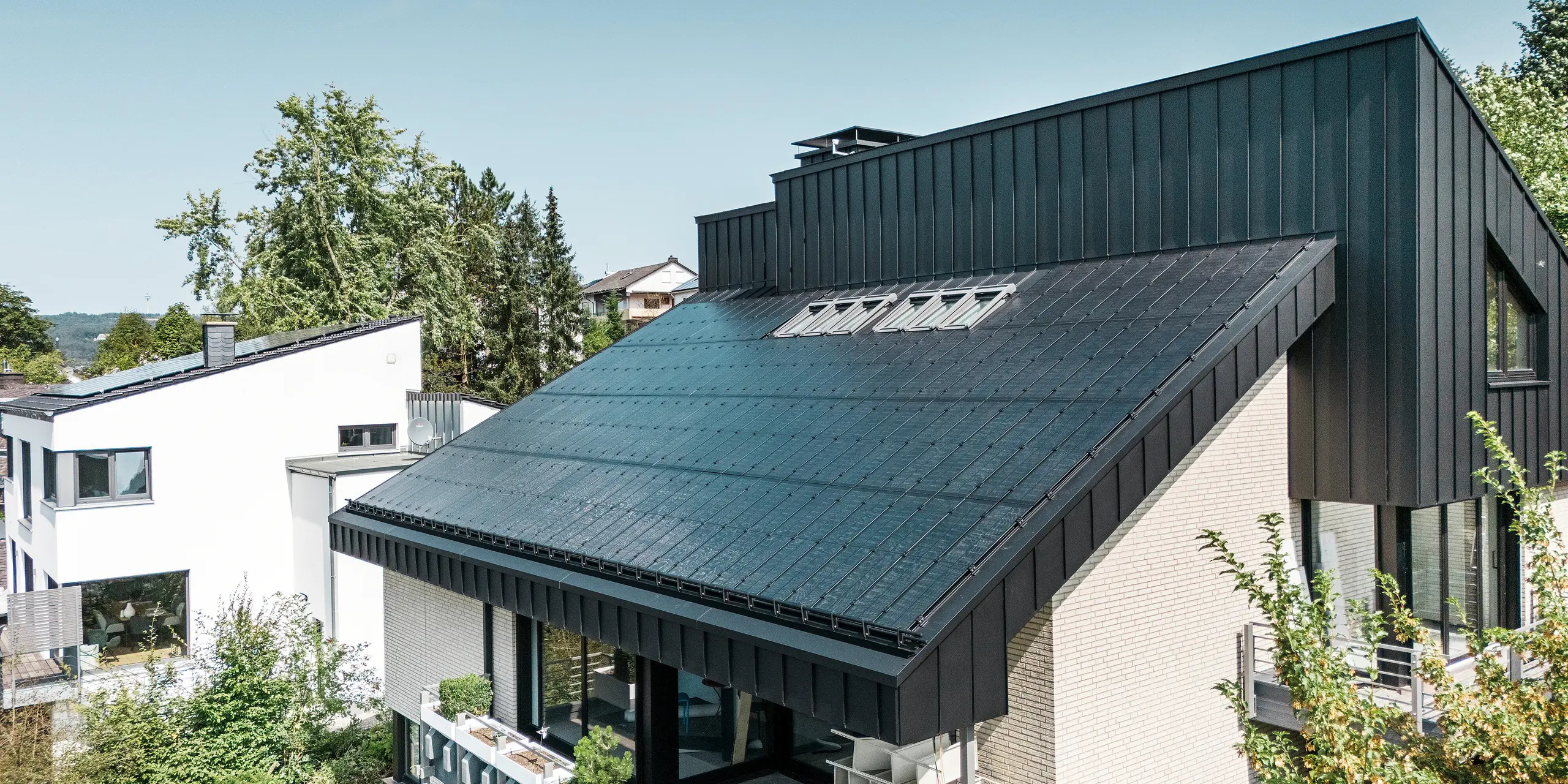 Architettura moderna di una casa unifamiliare a Meschede, in Germania, con un tetto in alluminio PREFALZ in P.10 nero e moduli fotovoltaici vetro/vetro integrati. Il tetto a una falda sfalsato combina un design senza tempo con una tecnologia fotovoltaica sostenibile. Gli innovativi moduli fotovoltaici PREFALZ forniscono una potenza di 18 kWp. I moduli senza cornice e l'elegante rivestimento in alluminio di facciata e tetto formano un'unità estetica e funzionale che si integra perfettamente nel verde ambiente residenziale.