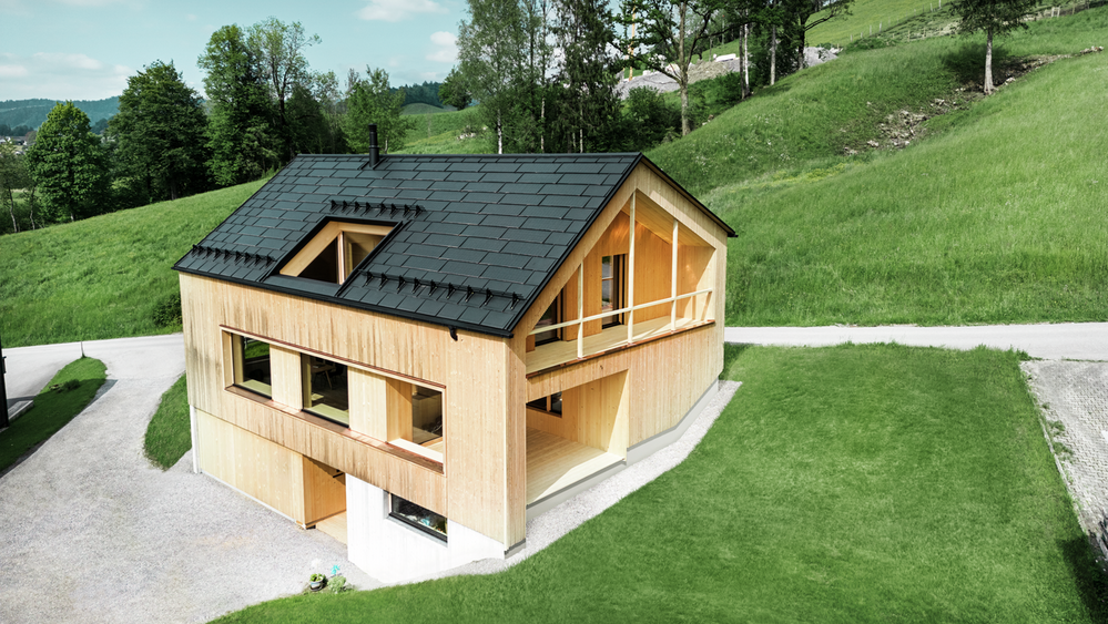 Ein modernes, zweistöckiges Haus mit einer Fassade aus hellem Holz steht in einer idyllischen, grünen Landschaft. Das schräge Dach des Hauses ist mit schwarzen PREFA Solardachplatten und R.16 Dachplatten versehen. Vor dem Haus befindet sich ein gepflegter Rasen, und eine geschwungene Schotterstraße führt zum Haus. Im Hintergrund erstrecken sich sanfte grüne Hügel mit Bäumen.