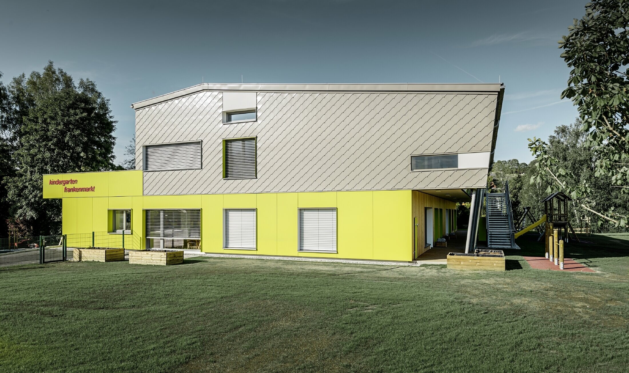 La scuola materna è stata in parte rivestita con la scaglia 29 PREFA in bronzo, anche questa parte dell'edificio è priva di sporto del tetto. Il resto della facciata è stato rivestito con pannelli di colore giallo.